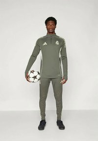 Chándal deportivo en verde oliva con acentos blancos, con un collar con cremallera. Sosteniendo un balón de fútbol, zapatillas negras, diseño ajustado.