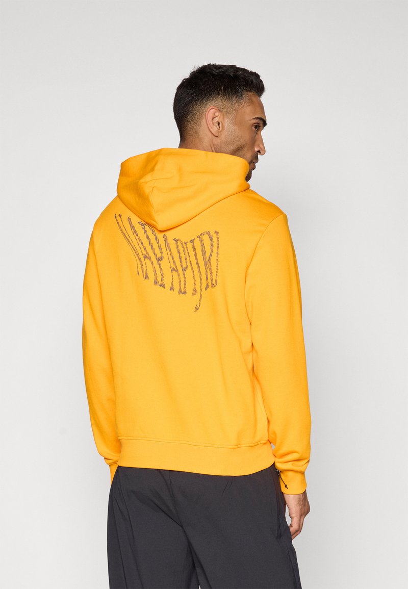 Mann, der seitlich steht, trägt einen leuchtend orangefarbenen Hoodie mit gewelltem schwarzem Text auf der Rückseite und dunkler Hose vor einem schlichten Hintergrund.