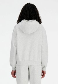 Felpa grigia con cappuccio, dal taglio rilassato, con polsini e fondo a costine; texture morbida e minimalismo nei dettagli. Indossata con pantaloni coordinati.