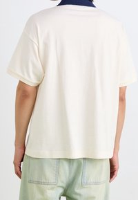 Personne portant une chemise crème ample à manches courtes avec un col bleu marine, associée à un pantalon vert clair, vue de dos.