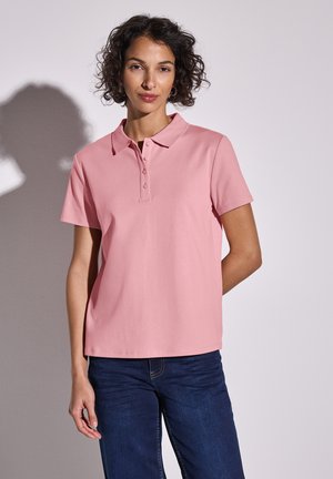 Lyserød polo shirt lavet af bomuld, med krave, korte ærmer og en tre-knap lukning. Sammen med mørkeblå jeans.