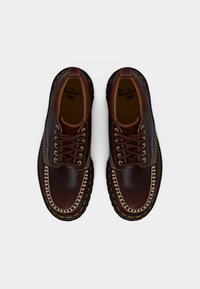 Ζευγάρι μοκασίνια από σκούρο καφέ δέρμα με κίτρινη ραφή και χοντρές μαύρες σόλες, σε θέα από πάνω σε λευκό φόντο.