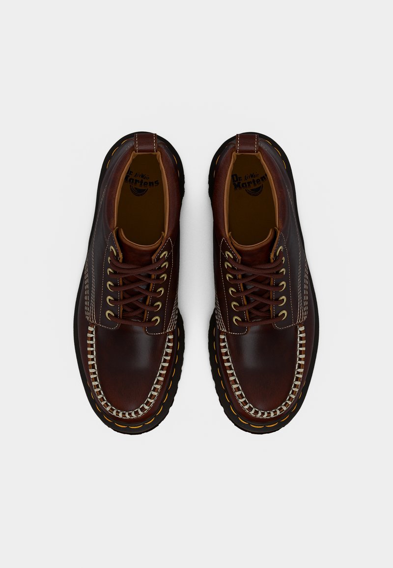 Een paar donkerbruine leren moc-toe schoenen met gele stiksels en dikke zwarte zolen, bekeken vanaf bovenaf tegen een witte achtergrond.