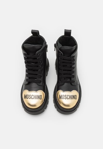 Las botas de tobillo de cuero negro presentan una tapa de punta en forma de corazón dorado con el logo "MOSCHINO", cordones robustos y cierre de cremallera lateral.