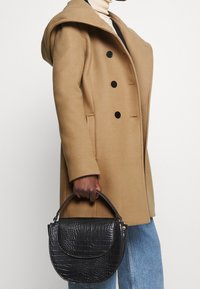 Person iført en kamelfarvet frakke og lyseblå jeans holder en sort håndtaske med krokodillemønster og top-håndtag foran en hvid baggrund.