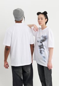 Camisetas blancas de gran tamaño, una con un gráfico de mariposas y un rostro, combinadas con jeans oscuros. Modelo llevando un gorro de punto gris.