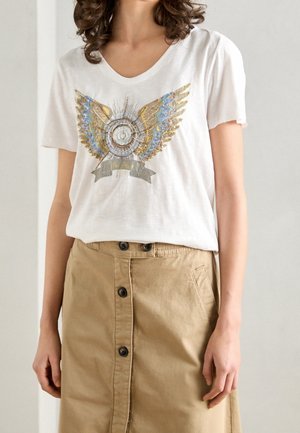 Kvinde iført en hvid T-shirt med et gyldent og blåt vinget emblem og en høj talje beige nederdel med knapper foran.