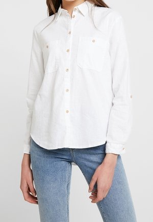 Femme portant une chemise blanche à manches longues boutonnée avec deux poches poitrine et un jean bleu clair sur un fond uni.