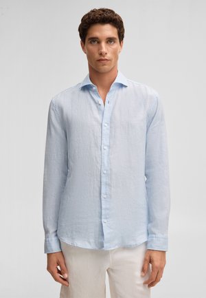 Mann trägt ein hellblaues, langärmliges, geknöpftes Hemd und weiße Hose, steht vor einem schlichten grauen Hintergrund.