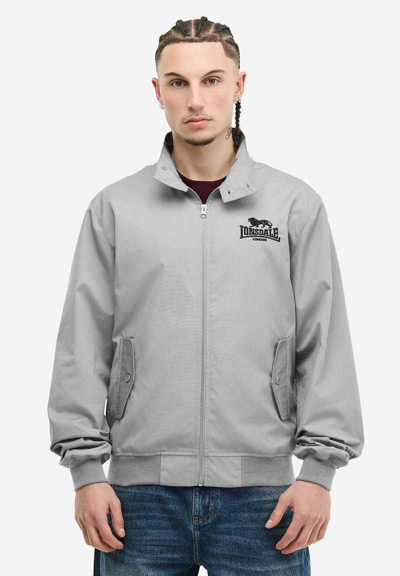 Jeune homme aux cheveux tressés portant une veste zippée gris clair avec le logo Lonsdale London et un jean bleu, debout face à l'avant.