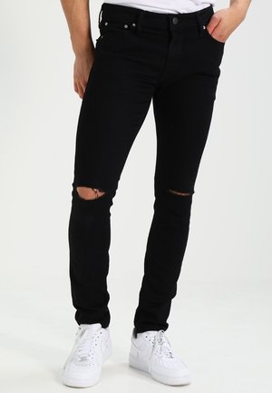 Jeans negros ajustados con rodilleras desgastadas, confeccionados en denim. Combinados con zapatillas blancas. Presenta bolsillos estándar y detalles metálicos en plateado.