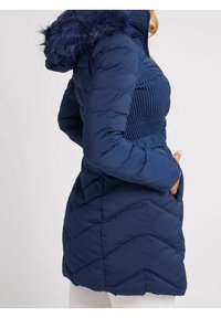 Mujer vestida con un abrigo de invierno acolchado de color azul marino, con una sección media texturizada y una capucha con piel sintética, con las manos en los bolsillos, mostrada de lado.