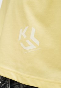 Κίτρινο ύφασμα με λευκό λογότυπο Karl Lagerfeld Jeans τυπωμένο κοντά στο τελείωμα, φορεμένο πάνω από μαύρο και γκρίζο τζιν.