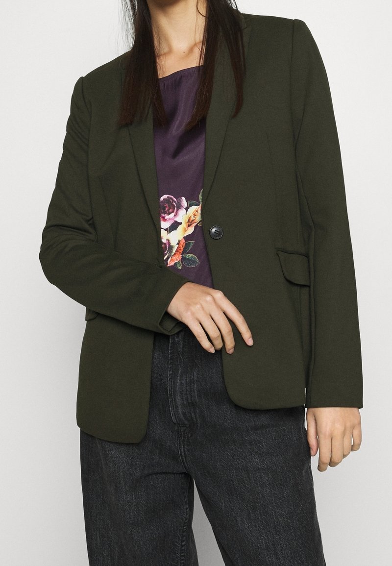 Mujer con un blazer verde oscuro sobre una blusa floral morada y pantalones de mezclilla negros, con una mano tocando el área de los botones del blazer.