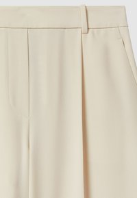 Beige maßgeschneiderte Hosen aus strukturiertem Stoff, mit Falten und seitlichen Taschen. Der Taillenbund ist mit einer Gürtelschlaufe versehen.