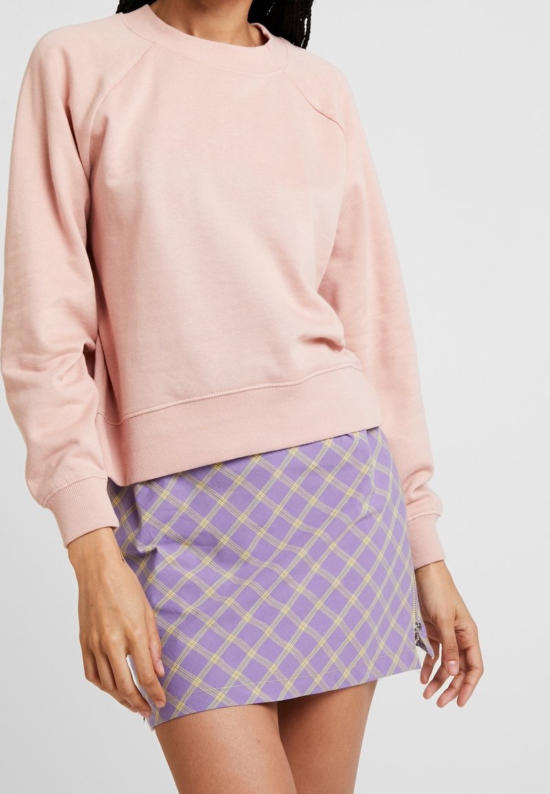 Personne portant un sweat-shirt rose clair à manches longues et une mini-jupe violette à motif à carreaux jaunes sur un fond uni.