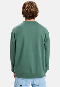 Zelená mikina s volným střihem, kulatým výstřihem a žebrovanými manžetami. Měkká textilie a minimalistické detaily na zadní straně.