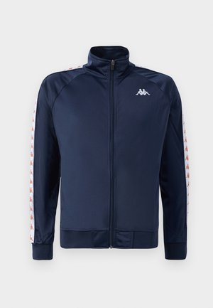 Navy blue athletic jacket with high collar, full zip, ribbed cuffs, and contrasting white side stripes featuring a repeated logo pattern.