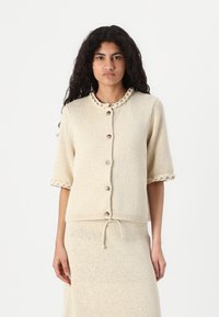 Cardigan à manches courtes en tricot de couleur beige clair, avec un tissu texturé, un col décoratif en tresse et une fermeture à boutons.