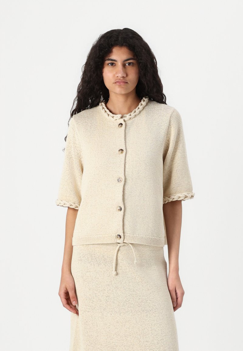 Cardigan à manches courtes en tricot de couleur beige clair, avec un tissu texturé, un col décoratif en tresse et une fermeture à boutons.