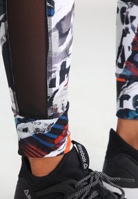 Piernas inferiores vestidas con zapatillas negras Reebok y leggings multicolores con paneles de malla negra y patrones abstractos.