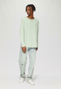 Jasnozielona koszula henley z długim rękawem, okrągłym dekoltem i trzema guzikami, zestawiona z jasnoniebieskimi przetartymi dżinsami i białymi trampkami.