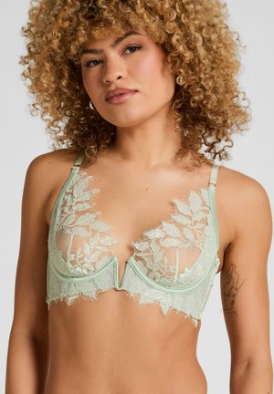 Vrouw met krullend blond haar, gekleed in een lichtgroene kanten beha met bladpatronen en verstelbare bandjes, tegen een effen achtergrond.