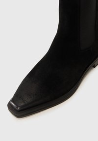 Bottine en suede noir à bout pointu, dotée d'un panneau latéral élastiqué et d'une semelle en cuir lisse, mettant en avant un design élégant et moderne.