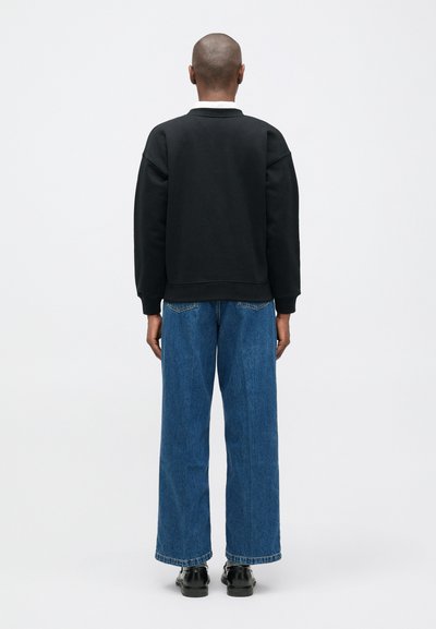 Persona de pie de espaldas, vistiendo un suéter negro, jeans anchos azules y zapatos negros, contra un fondo blanco liso.