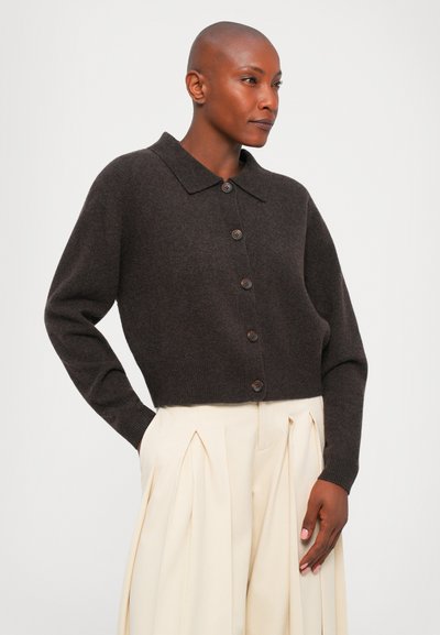 Personne avec la tête rasée portant un gilet boutonné marron foncé et un pantalon crème plissé taille haute, se tenant avec une main dans sa poche.