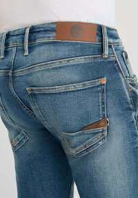 Los pantalones vaqueros de mezclilla azul tienen una textura ligeramente desgastada, cinco bolsillos y una etiqueta de cuero marrón en la cintura con un logotipo circular.