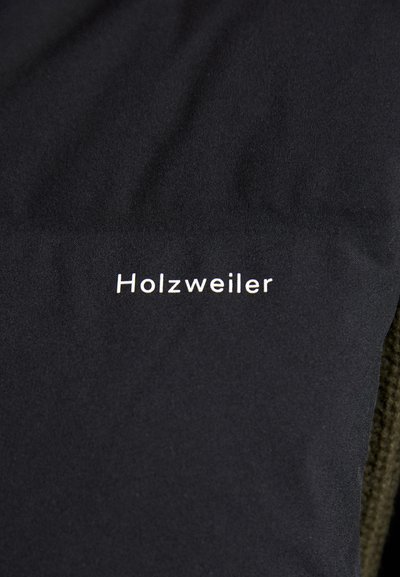 Svart jakke med en jevn tekstur med "Holzweiler"-logo i hvitt, som kontrasterer med et grønt, teksturert stoff på den ene siden.