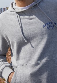 Šedá mikina s kapucí z měkké tkaniny. Má přední kapsu, kapuci se šňůrkou na stažení a modrý nápis "ATHLETIC Club" s vavřínovými akcenty.