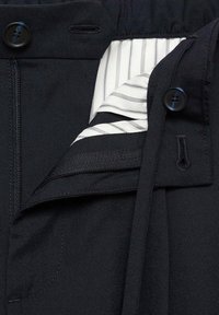 Ceinture de pantalon noire avec fermeture éclair, bouton et doublure intérieure rayée blanche partiellement visible sous le rabat.