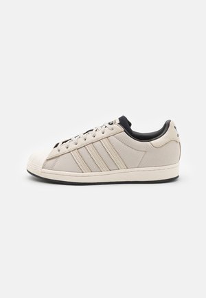Beżowy sneaker Adidas z trzema bocznymi paskami, designem z wzmocnionym noskiem, czarną wyściółką wewnętrzną i czarną podeszwą, pokazany z boku na białym tle.