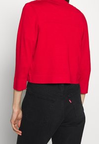 Chaqueta roja corta con mangas tres cuartos, hecha de tejido texturizado, combinada con jeans negros que presentan costuras sutiles y detalle de logotipo.