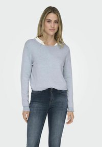 Hellblaues Sweatshirt mit langen Ärmeln und Rundhalsausschnitt, über einem weißen Hemdkragen getragen, kombiniert mit dunkelblauen Jeans.