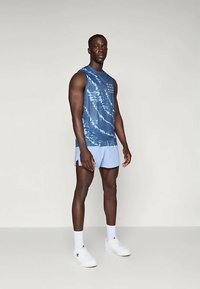 Men's blue tie-dye sleeveless shirt with a graphic design, paired with light blue shorts and white sneakers. Smooth fabric, athletic fit.