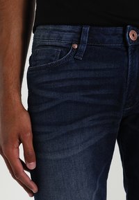 Mörkblå denimjeans med en subtil blekning, fem fickor och en kopparfärgad knappdetalj vid midjan.