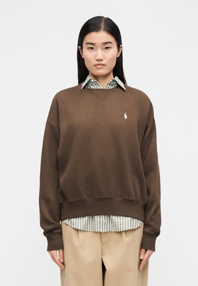 Ženska s ravnimi črnimi lasmi nosi rjavo majico čez zeleno-belo črtasto srajco in bež hlače, stoji ob preprostem ozadju.