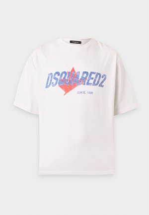 Uomo che indossa una t-shirt bianca Dsquared2, pantaloni beige, scarpe nere e berretto blu navy, in piedi davanti a uno sfondo uniforme.