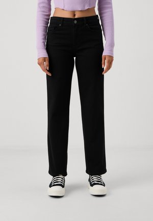 Kvinde iført sorte, højtaljede, lige jeans, sort-hvide sneakers og en cropped lavendel langærmet top, stående mod en ensfarvet baggrund.