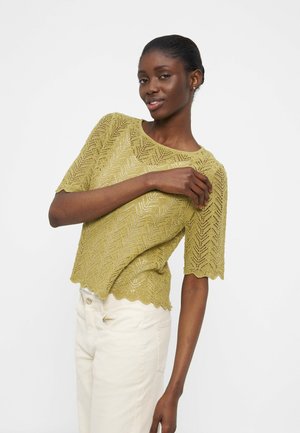 Kvinne iført en lys grønn kortermet genser med sikksakk-mønster og off-white høytlivsbukser, poserer med en arm krysset over brystet.