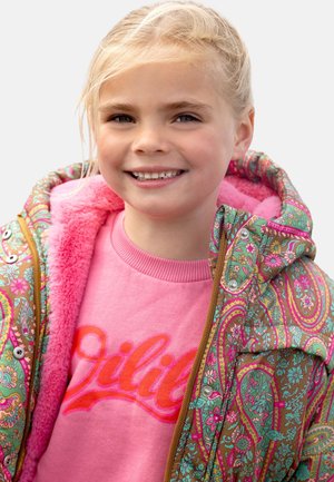 Kleurrijke patroonjas met een bloemen- en paisleypatroon in bruin, roze en groen, gevoerd met roze fleece, gedragen over een roze sweatshirt.