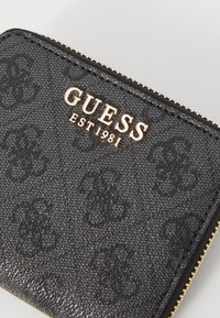 Crna sintetička novčanica s teksturiranim uzorcima i urezanim logotipima, zlatnim detaljima i zatvaračem na patentni zatvarač. Ispisano je "GUESS EST 1981".