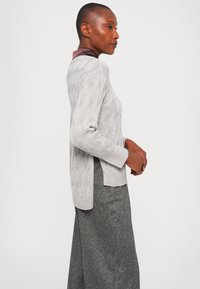 Woman in gray cable-knit sweater over a dark satin-collared shirt and loose gray pants, standing sideways against a plain background.