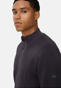 Mænds mørkegrå zip-up sweater med høj krave, tekstureret stof og et logo-patch på venstre ærme. Har et slankt design og subtile mønstre.