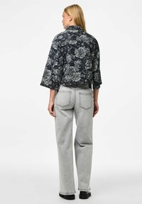 Vrouw in een zwart en grijs bloemenjacket met een cropped model en brede mouwen, draagt lichtgrijze hoogwaisted rechte jeans, staand met haar rug naar de camera toe.