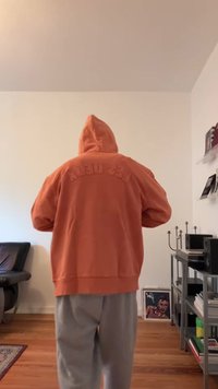 Eine Person mit orangefarbener Kapuzenjacke, die mit dem Rücken zur Kamera steht, in einem Raum mit Holzboden, schwarzem Stuhl und Metallschränken.
