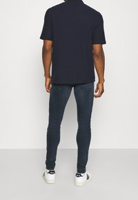 Polo de manga corta en color navy combinado con jeans ajustados de color azul oscuro y zapatillas blancas con detalles en negro, visto desde atrás.